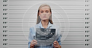 Mugshot of beautiful arrested woman holding sign for photo in police station. Crop view of young female criminal raising