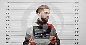 Mugshot of handsome guy with beard turning to sides while holding sign and posing for photo. Crop view of young criminal