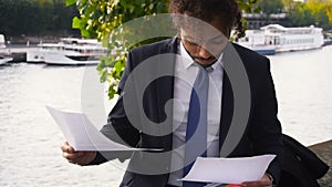 Mulatto student reading coursework near river and working with laptop.