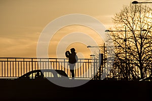Silhouette of woman taking a photography on bridge with smartphone in hands by sunset