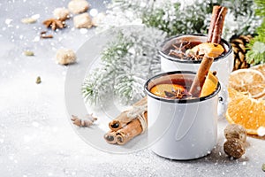 Mulled wine in white metal mugs with cinnamon, spices and orange with fir tree and christmas lights - traditional drink on winter