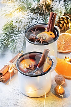 Mulled wine in white metal mugs with cinnamon, spices and orange with fir tree and christmas lights - traditional drink on winter