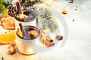 Mulled wine in white metal mugs with cinnamon, spices and orange with fir tree and christmas lights - traditional drink on winter