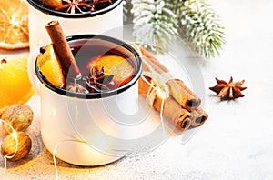 Mulled wine in white metal mugs with cinnamon, spices and orange with fir tree and christmas lights - traditional drink on winter