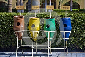 Multi-colored clay bins for separate waste collection. Urns with signs are lined up in row, against background of green bushes.