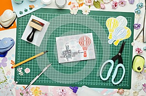 Multi-colored paper, homemade postcard and scrapbooking tools and materials on green mat for cutting