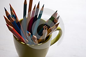Multi-colored pencils in green cup on wooden white table background - front viewn