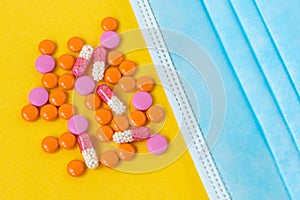 Multi-colored pills and a blue disposable mask on a bright yellow background, top view. Medication for the disease. Drugs from cor