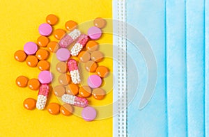 Multi-colored pills and a blue disposable mask on a bright yellow background, top view. Medication for the disease. Drugs from cor