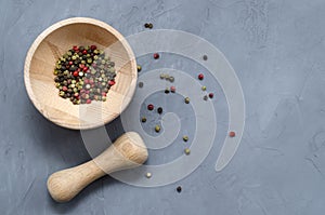 Multi-colored red green black peppercorns in a wooden mortar with a pestle and scattered around on a ultimate gray