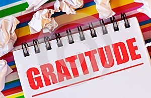 On a multi-colored striped background there are white pieces of paper and a white notebook with the inscription in red GRATITUDE