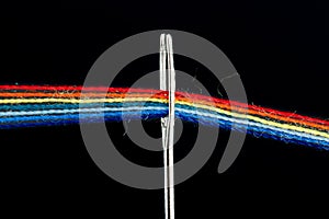 Multi-colored threads for sewing in the form of a rainbow pass through an antique needle on a black background