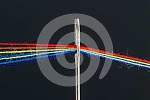 Multi-colored threads for sewing in the form of a rainbow pass through an antique needle on a black background