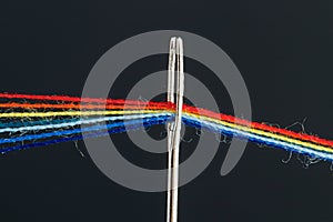 Multi-colored threads for sewing in the form of a rainbow pass through an antique needle on a black background