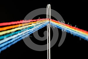 Multi-colored threads for sewing in the form of a rainbow pass through an antique needle on a black background