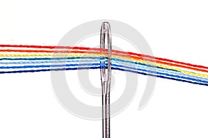 Multi-colored threads for sewing in the form of a rainbow pass through an antique needle on a white background