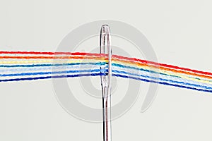 Multi-colored threads for sewing in the form of a rainbow pass through an antique needle on a white background