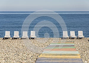 Multi-colored wooden walkway leading to white loungers standing on a pebble beach next to the shore