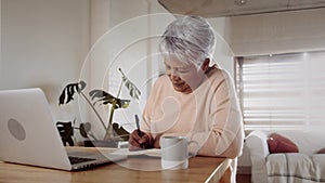 Multi-cultural elderly female searching online using laptop, sitting at kitchen counter. Pen and paper making notes