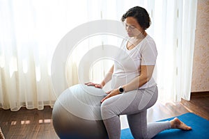 Multi ethnic adult pregnant woman exercising with a fit ball at home, for wellness and wellbeing in pregnancy time