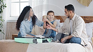 Multi-ethnic family multiracial young parents with little daughter girl child sitting on bed in bedroom packing things