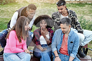 Multi-ethnic friends having fun and laughing while using a mobile phone sitting on a bench outdoors.