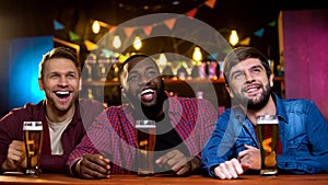 Multi-ethnic friends smiling watching comedy show on big screen in beer pub, fun