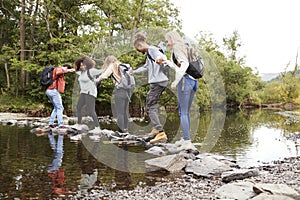Multi ethnic group of five young adult friends hold hands balancing on rocks to cross a stream during a hike