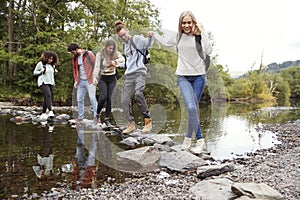 Multi ethnic group of five young adult friends hold hands walking on rocks to cross a stream during a hike