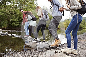 Multi ethnic group of five young adult friends hold hands walking on rocks to cross a stream during a hike, close up, back view