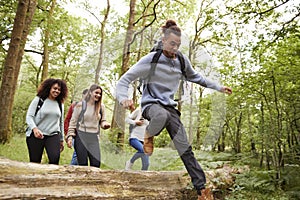 Multi ethnic group of five young adult friends running in a forest and jumping over fallen tree during a hike