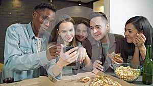 Multi-ethnic group of friends looking at smartphone talking laughing and eating pizza and snacks at a bar or at home