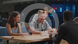 A multi-ethnic group of friends sits at a table together in a bowling alley and talks. Friends met together in a bowling