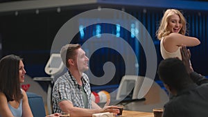 A multi-ethnic group of friends sits at a table together in a bowling alley and talks give five joyful blonde woman