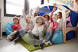 A multi-ethnic group of infant school children sitting on bean bags in a comfortable corner of the classroom, raising their hands