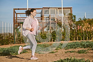 Multi ethnic middle aged sporty woman in gray leggings and pink hoodie jogging on the beach at sunset