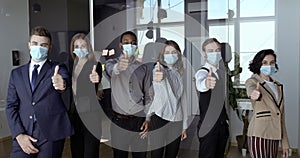Multi ethnic team of young ambitious employees wearing face masks, posing positive with thumb up gesture, show support
