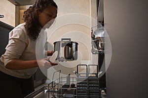 Multi ethnic woman, housewife unloading a clean stainless saucepan from a dishwasher. Modern kitchen appliances