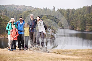 Multi generation family stand embracing by a lake, smiling to camera, front view, Lake District, UK