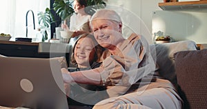Multi generational family bonding. Happy senior 60s grandmother using laptop smiling with teen girl at cozy home couch.