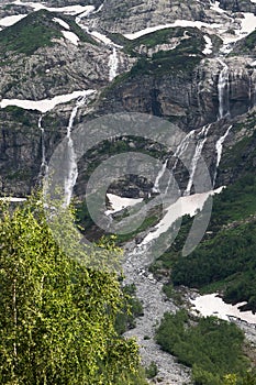 A multi-jet alpine waterfall on a high cliff with a green forest at the foot with glimpses of snow. The concept of a