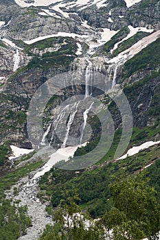 A multi-jet alpine waterfall on a high cliff with a green forest at the foot with glimpses of snow. The concept of a