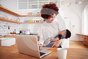 Multi-Tasking Mother Holds Sleeping Baby Son And Works On Laptop Computer In Kitchen