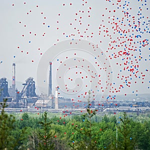 Multicolored balloons in the sky against the background of the pipes of a large metallurgical plant. Contrasts of nature, industry