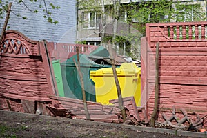 Multicolored garbage containers are visible through the broken wall of the place for household waste