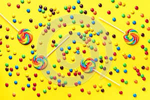 Multicolored lollipop, rainbow colorful candies on yellow background. Coated chocolate sweet pieces texture. Top view. Flat lay.