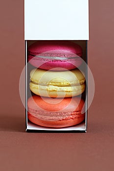 Multicolored macarons in a cardboard box with an open lid standing upright on a brown background. Closeup