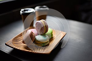 Multicolored macarons in a wooden plate and two cups with coffee in dark colors