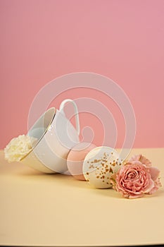 Multicolored macaroons cake are placed in a saucer and a cup along with buds of a carnation and roses on a yellow and pink