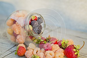 Multicolored raspberries in a glass jar with strawberries on background